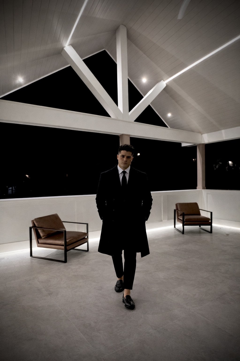Him — a man in a dark coat on the pergola after dark, the night behind him.